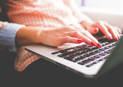 En person sitter med laptop i fanget og skriver. Personen har lange røde negler og rutete skjorte.  Bildet er ett utsnitt av tastaturet til pc-en og nedre del av overkroppen. - Klikk for stort bilde