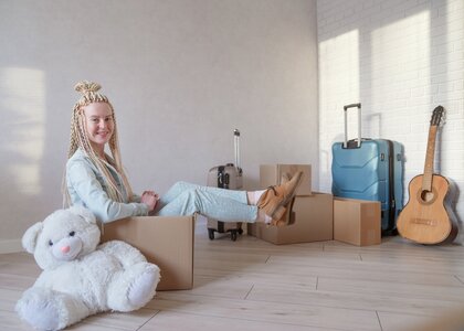 Ung kvinne som sitter i en pappeske med diverse ting rundt seg - bamse, gitar, koffert  og flere pappesker - Klikk for stort bilde